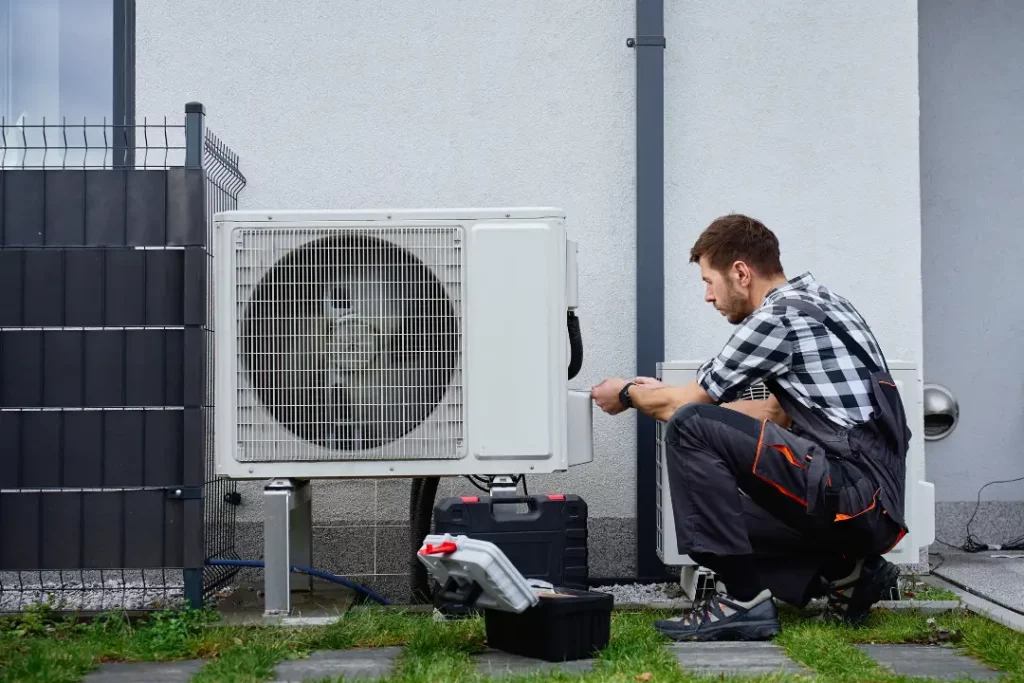 Fachmann prüft kniend eine Wärmepumpe, welche vor einer Hauswand steht. 