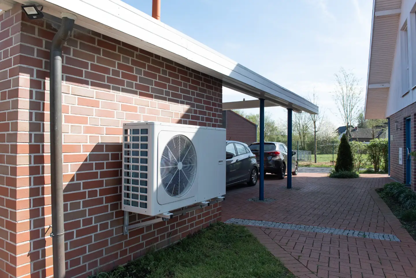An einer Garage gegenüber eines Hauses steht eine Wärmepumpe. Im Hintergrund sieht man zwei Autos vor der Garage.